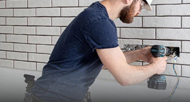Electrician using a cordless drill to install wiring in a tiled wall, wearing a hard hat and safety glasses for protection.