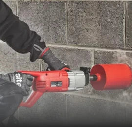Worker using a red core drilling machine with a large diamond core bit to drill a circular hole into a concrete block wall.