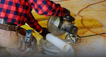 Plumber in flannel shirt using a miter saw to cut a white PVC pipe on a workshop floor, surrounded by wooden planks and tools.