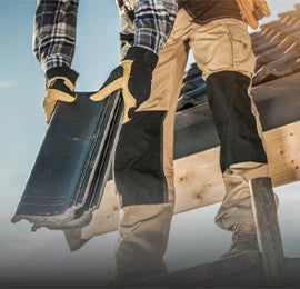 Roofer wearing gloves and workwear placing black roofing tiles on a wooden roof frame, standing securely on a ladder.