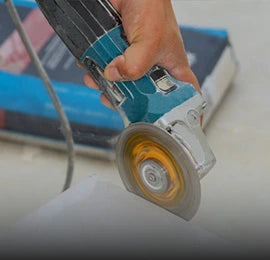 A close-up of a hand using an electric angle grinder to cut a tile, with motion blur on the spinning blade and tools in the background.