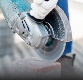 A gloved hand using a large electric angle grinder to cut a concrete block, with visible motion blur on the spinning cutting disc.